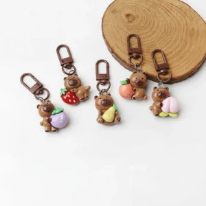 A collection of five cute resin capybara bag charms arranged in a row, each holding a different colorful fruit including a pear, persimmon, strawberry, peach, and mangosteen.