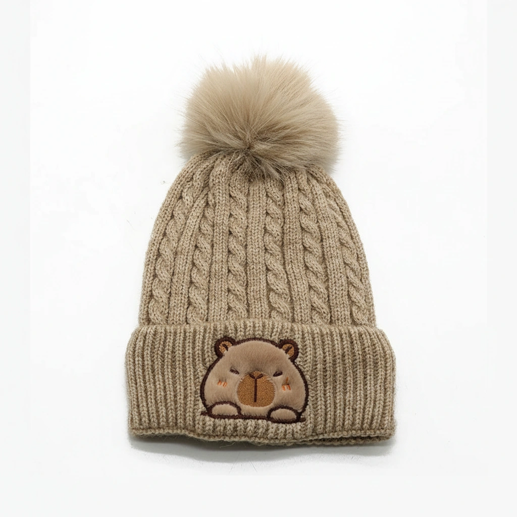 Beige cable knit winter hat for kids featuring a large fluffy pom-pom and a fuzzy embroidered capybara patch on the cuff.