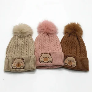 3 colors collection of cable knit winter hat for kids featuring a large fluffy pom-pom and a fuzzy embroidered capybara patch on the cuff.