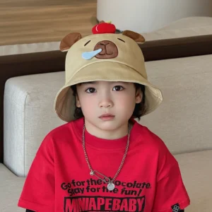 Toddler wearing a khaki cotton bucket hat featuring a 3D red apple on top and a sleeping capybara face with a blue bubble, looking at the camera.