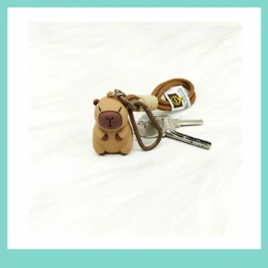 Close-up of a 3D PVC capybara keychain attached to a brown leather bag, featuring a utility rope strap and metal carabiner clip.
