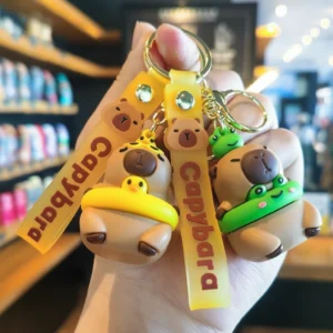 Close-up of a 3D cartoon capybara keychain made of PVC, wearing a green frog swim ring and holding a yellow strap with gold alloy hardware.