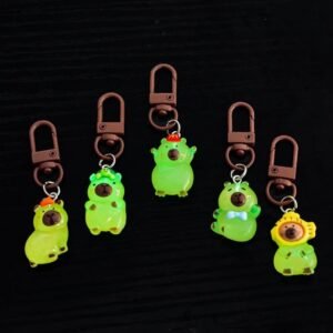 Set of 5 resin capybara bag charms shown in both daylight as translucent brown figures and in the dark glowing bright green, featuring designs with turtles, oranges, and hats.