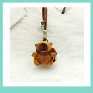 Front view of capybara plush pendant hanging by a brown woven rope strap, wearing a red and green scarf.