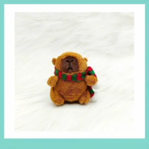A sitting capybara stuffed toy wearing a festive winter scarf with white pom-poms on a white background.