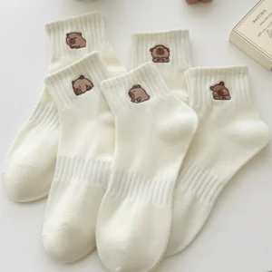 Five pairs of white ribbed crew socks arranged on a table, each featuring a different small embroidered brown capybara design on the cuff.